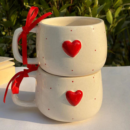Two ceramic mugs with red heart designs on a white surface with a green leafy background.