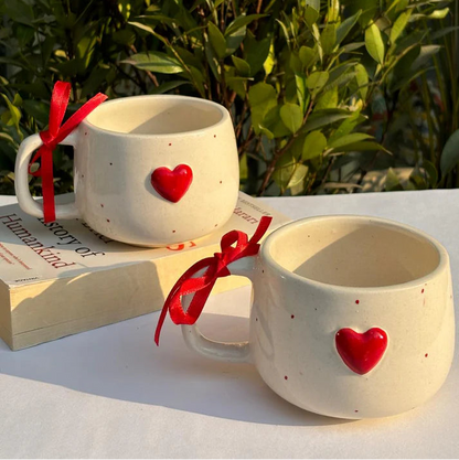 Two ceramic mugs with red heart designs on a white surface with greenery in the background.