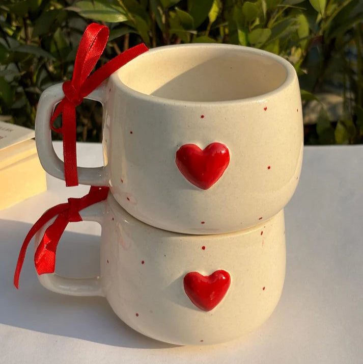 Two white mugs with red hearts and ribbons on a white surface with greenery in the background.