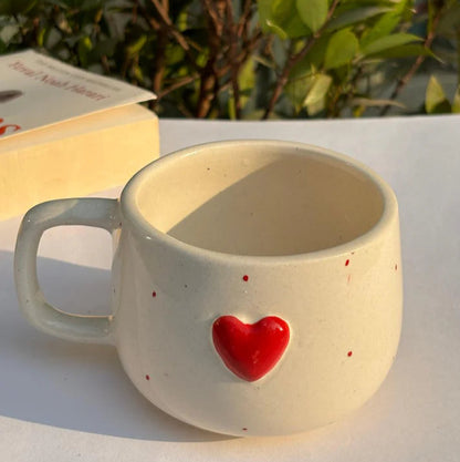 Ceramic mug with a red heart on a white surface with greenery in the background