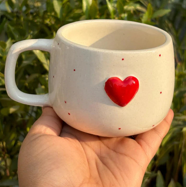 White mug with a red heart design held in front of green foliage