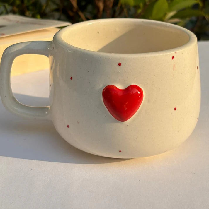 White ceramic mug with a red heart on a white surface with greenery in the background