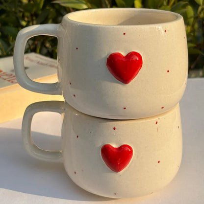 Two white ceramic mugs with red heart designs stacked on a reflective surface with greenery in the background.