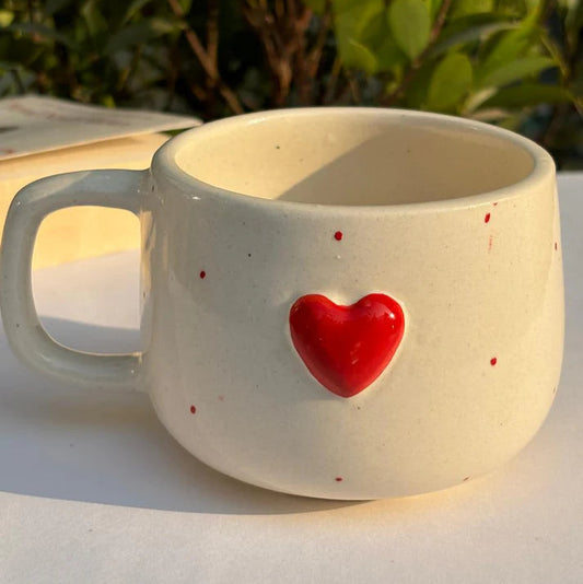 Ceramic mug with a red heart on a white surface with greenery in the background