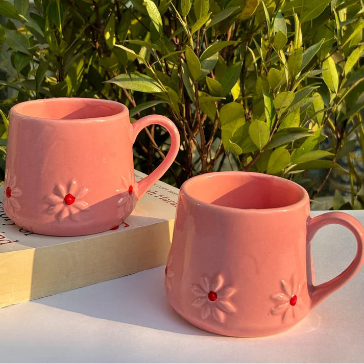 Two pink mugs with floral designs on a white surface with green foliage in the background