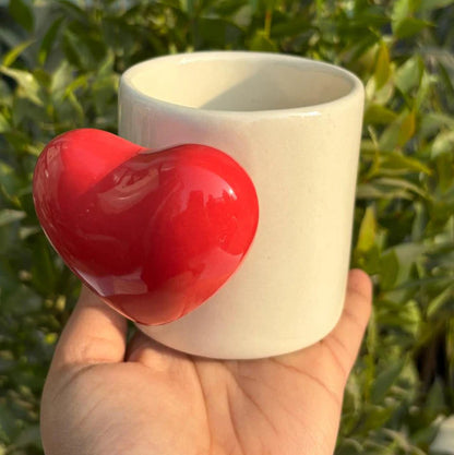 White ceramic cup with a red heart-shaped object held against a green leafy background