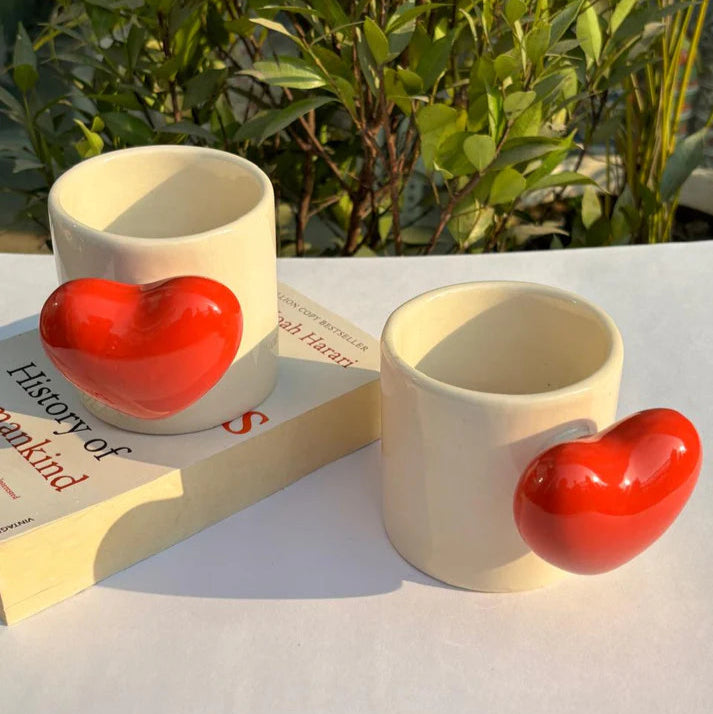 Two ceramic mugs with red heart designs on a white surface with greenery in the background.