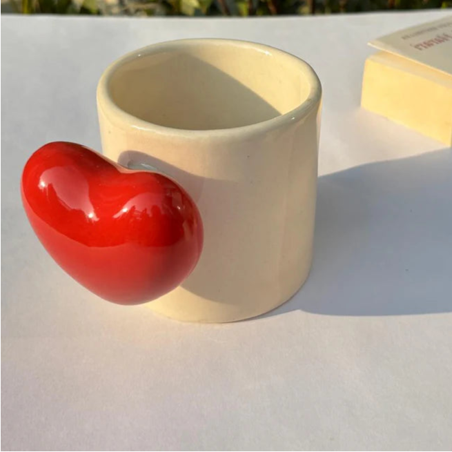Beige cylindrical container with a red heart-shaped object on a light surface with greenery in the background