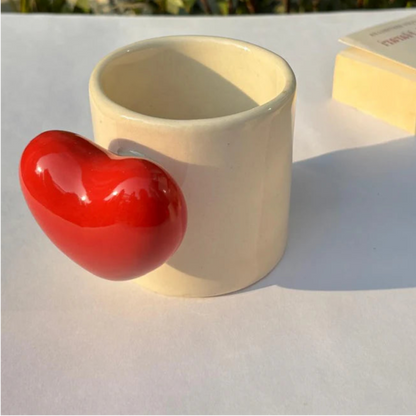 Beige cylindrical container with a red heart-shaped object on a light surface with greenery in the background