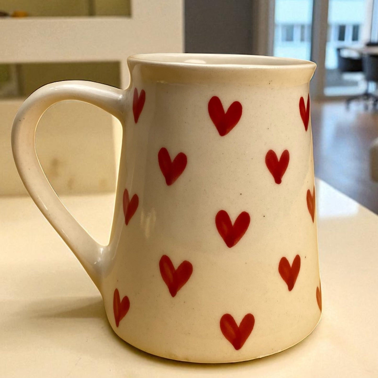 White mug with red heart patterns on a light-colored surface, possibly in a home setting.