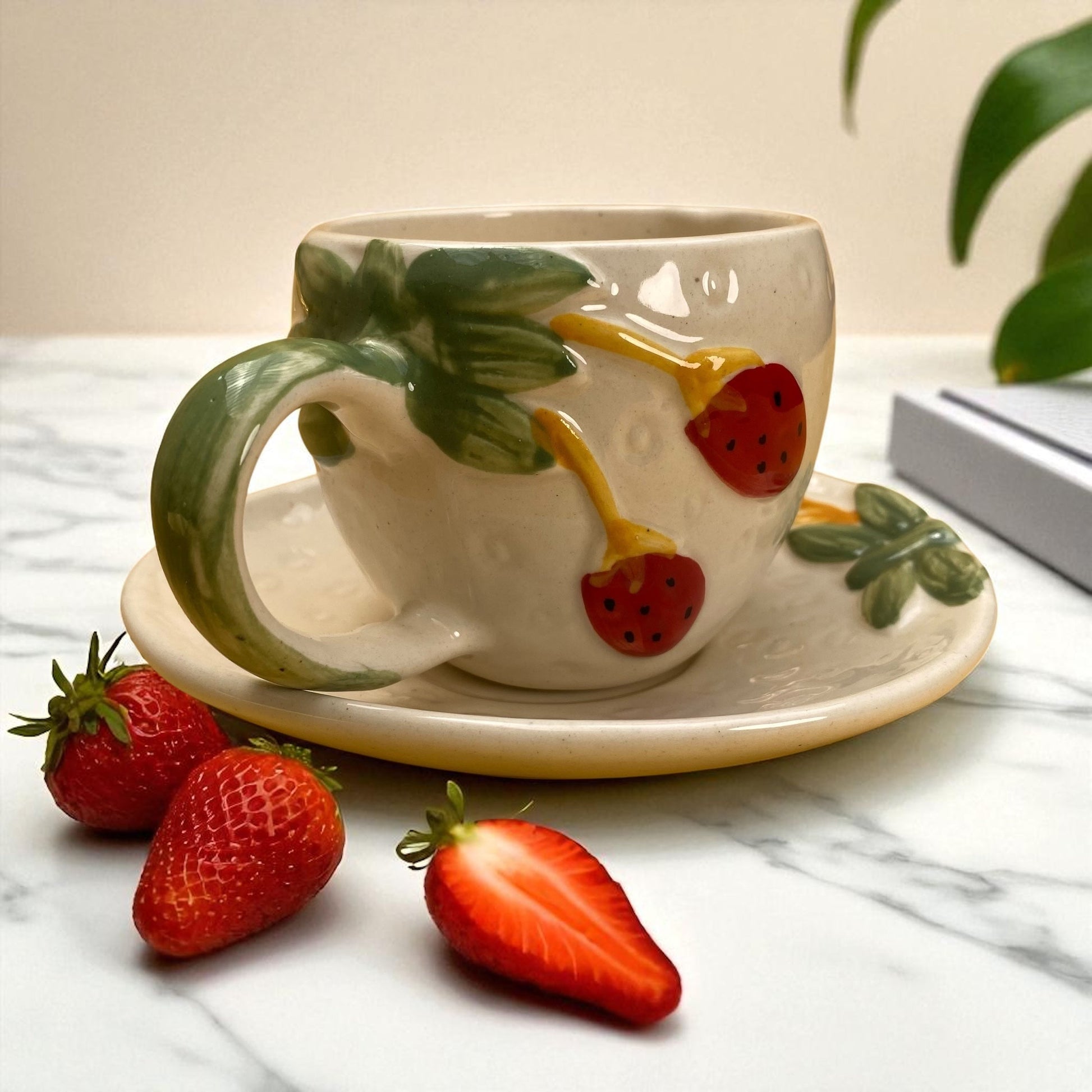 Ceramic mug with strawberry design on handle and saucer, surrounded by strawberries on a marble surface.