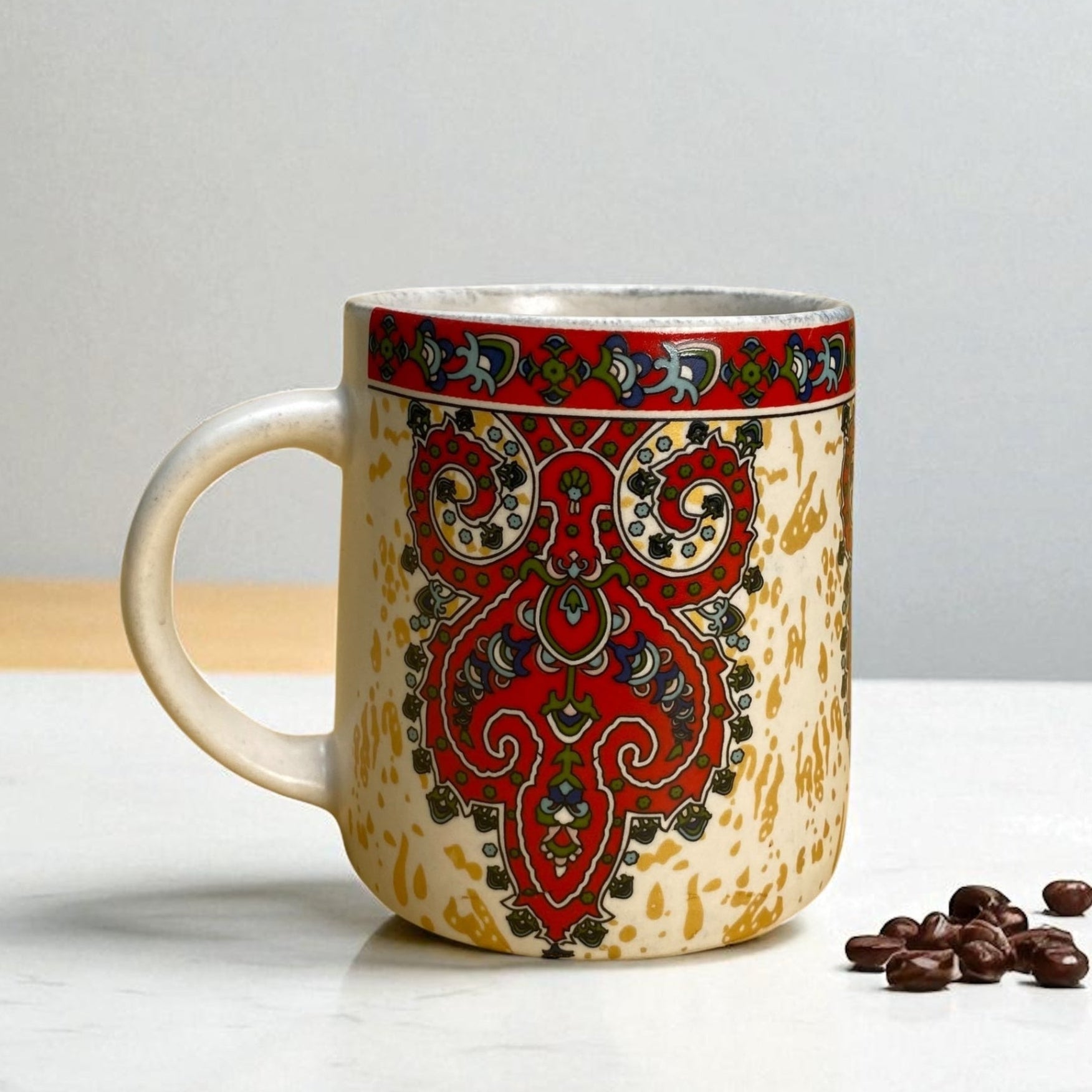 Decorative ceramic mug with red and green pattern on a white surface with coffee beans.