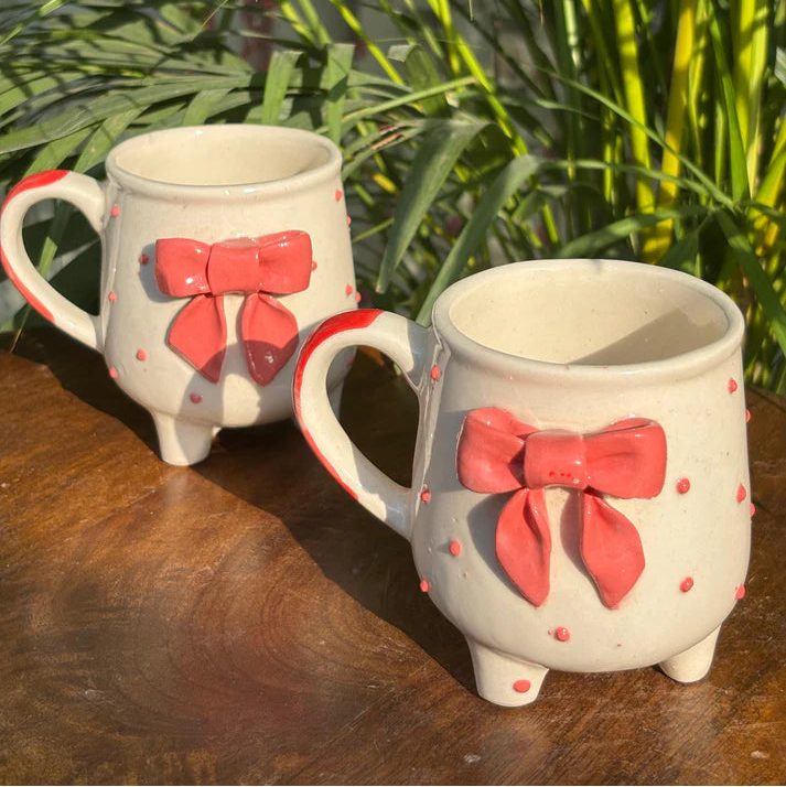 Two ceramic mugs with red bow designs on a wooden surface with green plants in the background.