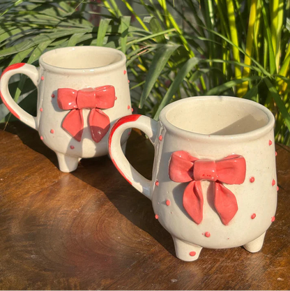 Two ceramic mugs with red bow designs on a wooden surface with green plants in the background.