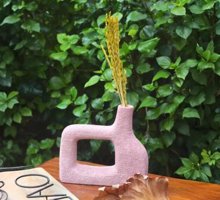 Pink vase with dried plants on a wooden surface against a green hedge background
