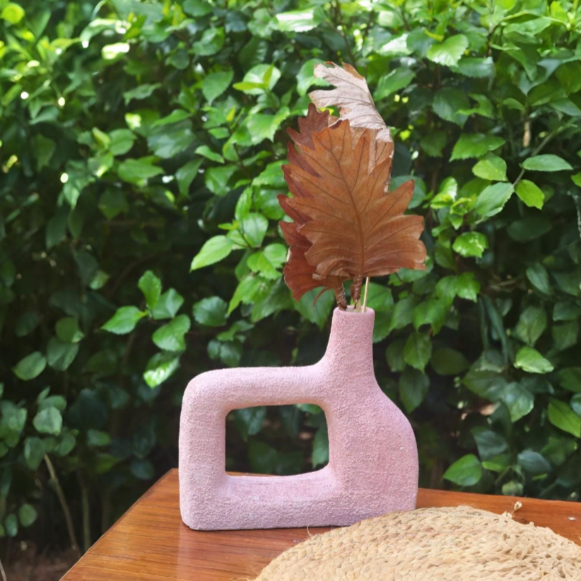 Pink textured vase with dried leaves against a green leafy background