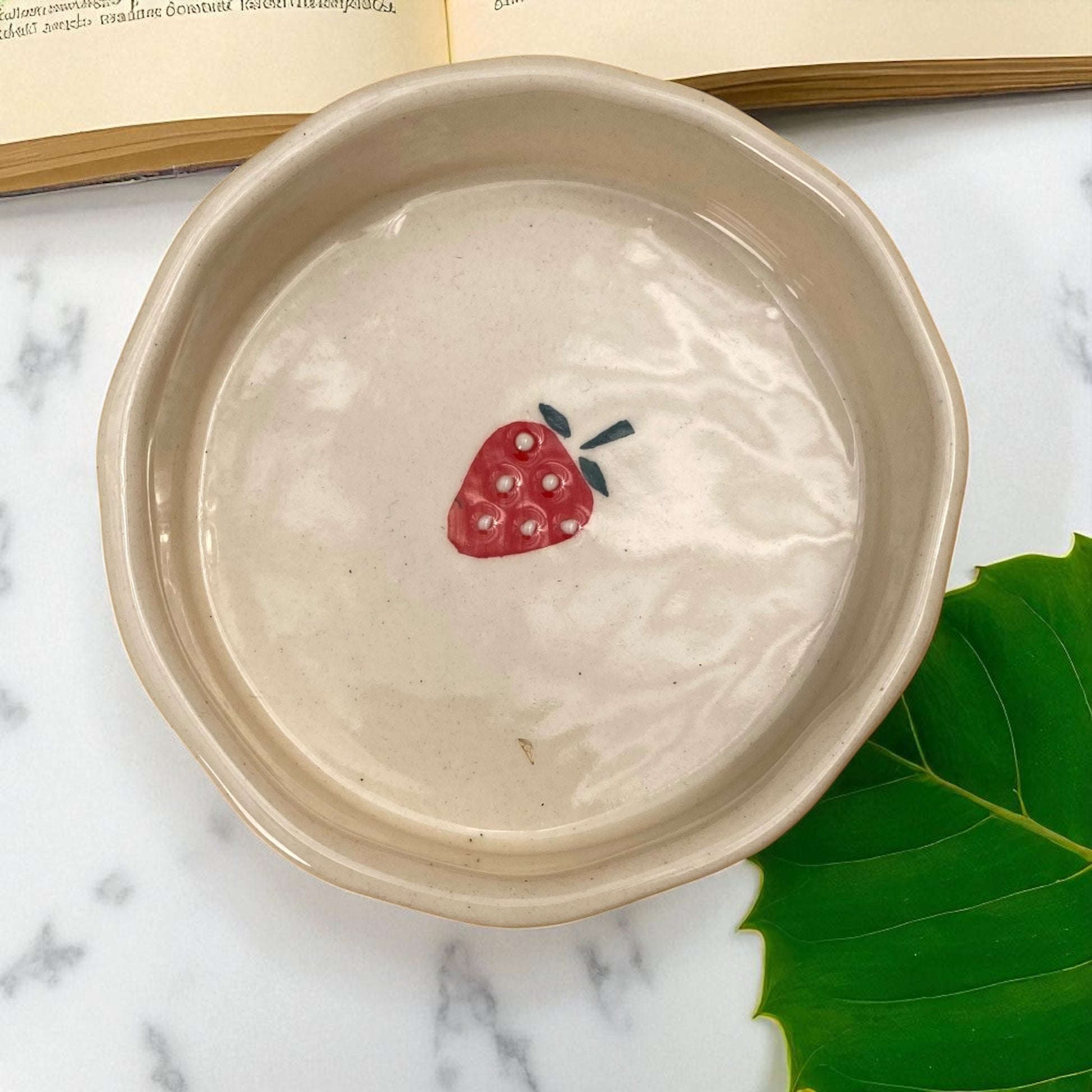 Ceramic bowl with a strawberry design on a marble surface with a green leaf.