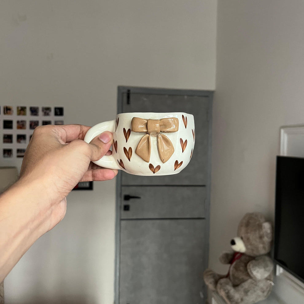 Person holding a decorative mug with a bow design in a room with a teddy bear and a television.